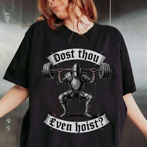 May include: Black t-shirt featuring a knight lifting a barbell in a squat position. The knight is clad in silver armor. The text "Dost thou" is above the knight, and "Even hoist?" is below.