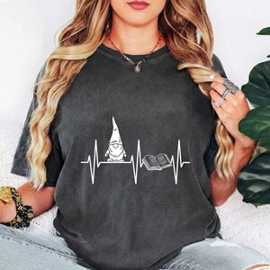 May include: Dark grey t-shirt with a white graphic of a gnome, an open book, and a heartbeat line. The gnome wears a tall, pointed hat. The shirt is made of a soft material.