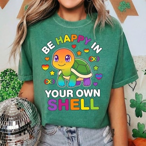 Może przedstawiać: Zielona koszulka z napisem "BE HAPPY IN YOUR OWN SHELL" i kolorową grafiką żółwia. Żółw ma tęczowy pancerz i jest otoczony sercami, gwiazdami i symbolami nieskończoności w tęczowych kolorach. Koszulka jest w stylu casual.