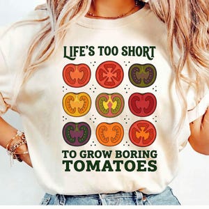 May include: Cream-colored t-shirt featuring a colorful tomato design. The design includes nine different tomato slices in various colors, with the text "LIFE'S TOO SHORT TO GROW BORING TOMATOES" in dark green.