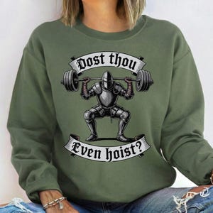 May include: Olive green sweatshirt with a graphic of a knight lifting a barbell. The knight is wearing full armor and is in a squat position. The text "Dost thou even hoist?" is displayed above and below the knight in a banner.