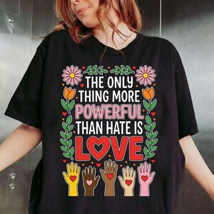 Puede incluir: Camiseta negra con el texto "THE ONLY THING MORE POWERFUL THAN HATE IS LOVE". El diseño incluye flores, hojas, corazones y manos de varios tonos de piel, cada una con un corazón. Una camiseta gráfica con un mensaje de amor y unidad.