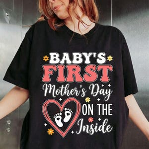 Può includere: T-shirt nero con la scritta "BABY'S FIRST Mother's Day ON THE Inside". Il design presenta un cuore con impronte di bambino e accenti floreali rosa, bianchi e gialli.