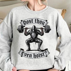 May include: A light gray sweatshirt featuring a graphic of a knight in armor lifting a barbell. The knight is in a squat position. The text "Dost thou" is above the knight, and "Even hoist?" is below.