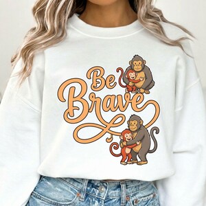 Op de afbeelding: Witte sweatshirt met de tekst "Be Brave" in oranje script. Het ontwerp toont twee sets cartoonapen die elkaar omarmen. De apen zijn bruin en oranje.