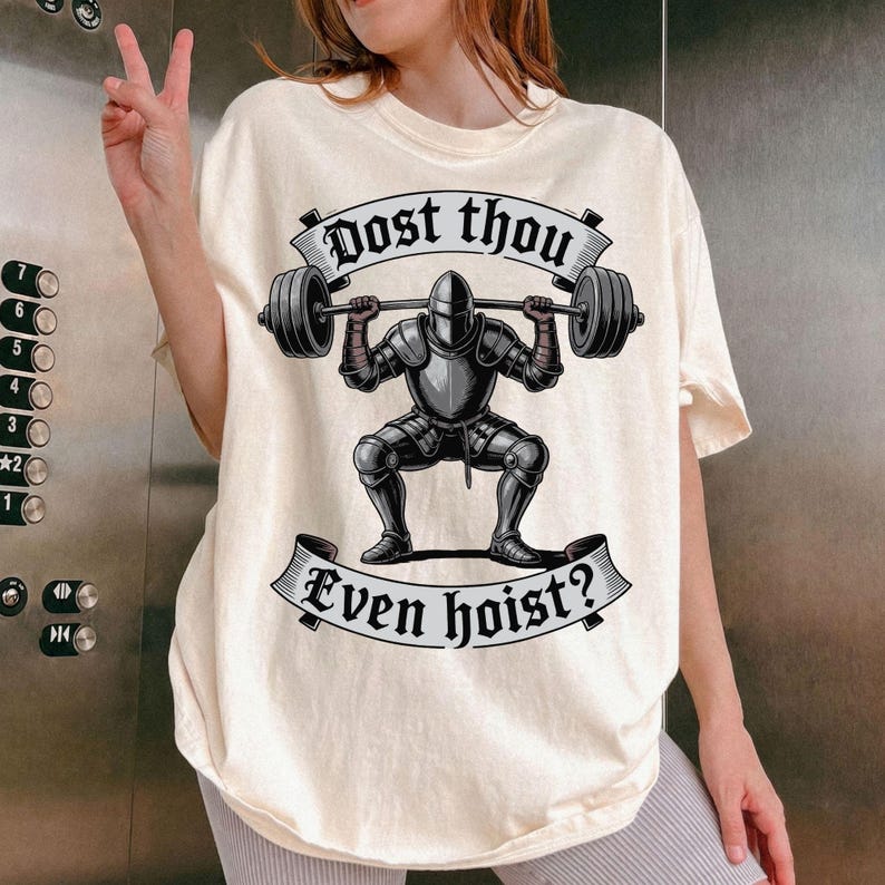May include: Cream-colored t-shirt featuring a graphic of a knight lifting weights. The knight is in full armor, and the text "Dost thou even hoist?" is displayed above and below the knight in a banner design.