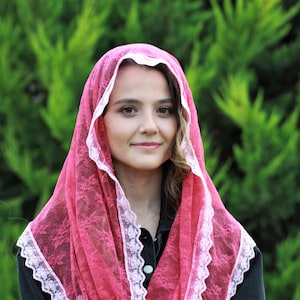 May include: A pink lace veil with white trim. The veil is worn over a person's head and shoulders.