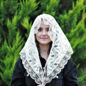 May include: A white lace shawl with a delicate floral pattern. The shawl is draped over a person's head and shoulders, with the edges of the shawl framing the person's face.