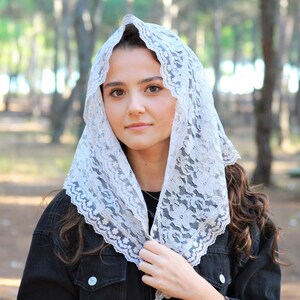 May include: A white lace head covering with a scalloped edge. The lace is delicate and intricate, with a floral pattern. The head covering is worn by a person with long brown hair.