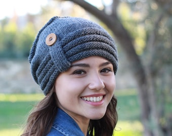 May include: A gray knitted beanie with a brown button on the side. The beanie is worn by a person with long brown hair and a denim jacket.