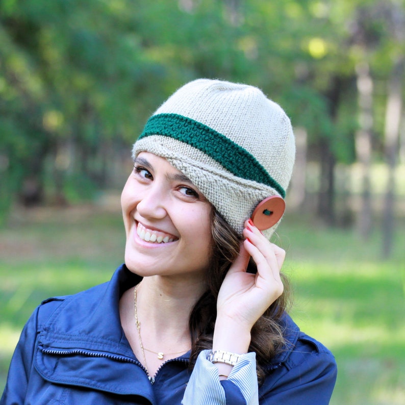 May include: A woman wearing a beige knitted hat with a green stripe and a brown button. The hat has a unique shape with a flap over the ear.