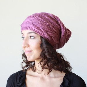 Pode incluir: Um gorro de malha, cor malva, com um design de malha entran&ccedil;ada. O chap&eacute;u &eacute; usado na cabe&ccedil;a, com a parte superior do chap&eacute;u ligeiramente ca&iacute;da. A pessoa est&aacute; vestindo um top preto.