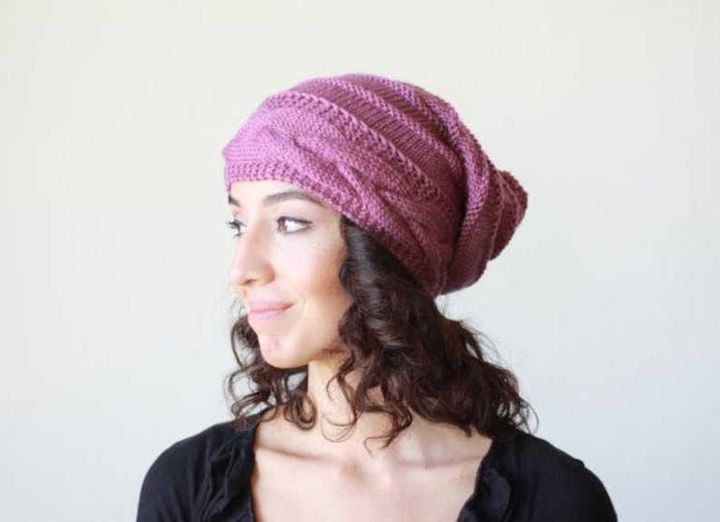 Pode incluir: Um gorro de malha, cor malva, com um design de malha entran&ccedil;ada. O chap&eacute;u &eacute; usado na cabe&ccedil;a, com a parte superior do chap&eacute;u ligeiramente ca&iacute;da. A pessoa est&aacute; vestindo um top preto.
