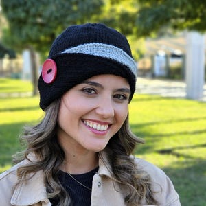 May include: A black knitted hat with a gray stripe and a red button. The hat has a cloche style and is worn by a person with long brown hair.