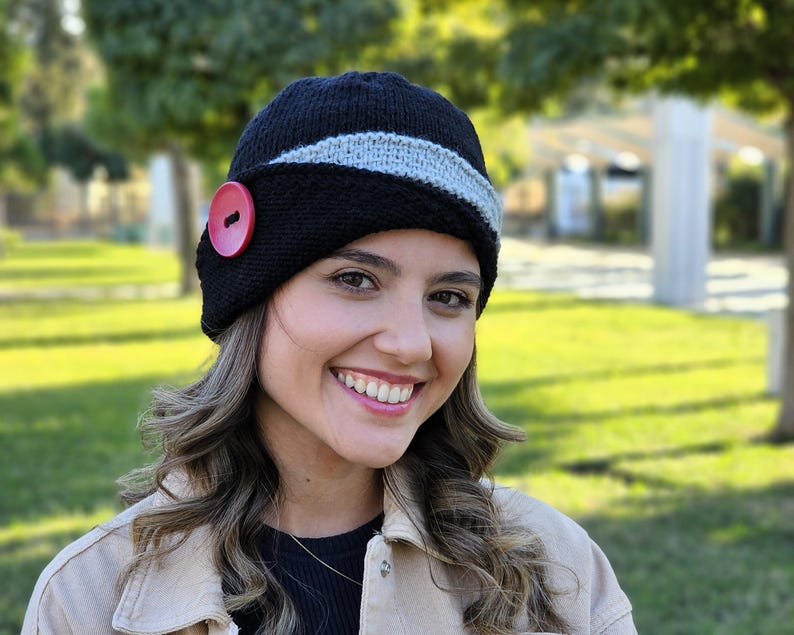 May include: A black knitted hat with a gray stripe and a red button. The hat has a cloche style and is worn by a person with long brown hair.