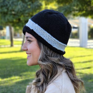 May include: A black knit beanie with a white stripe around the brim. The beanie is worn by a person with long brown hair.