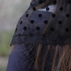 May include: Black sheer fabric with large velvet polka dots and a delicate lace trim. The fabric is draped over a dark blue garment, with long brown hair visible beneath the fabric.
