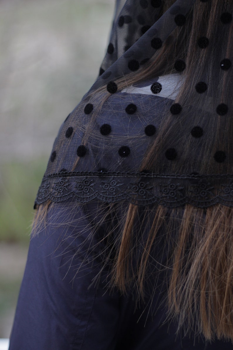 May include: Black sheer fabric with large velvet polka dots and a delicate lace trim. The fabric is draped over a dark blue garment, with long brown hair visible beneath the fabric.