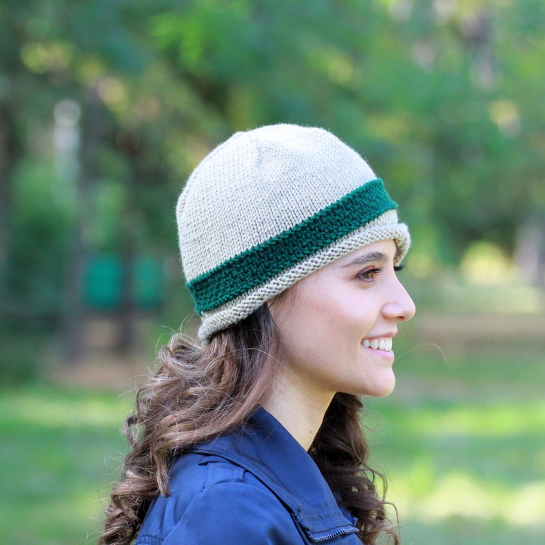 May include: A beige knitted beanie with a green stripe around the brim. The beanie is worn by a person with long brown hair.