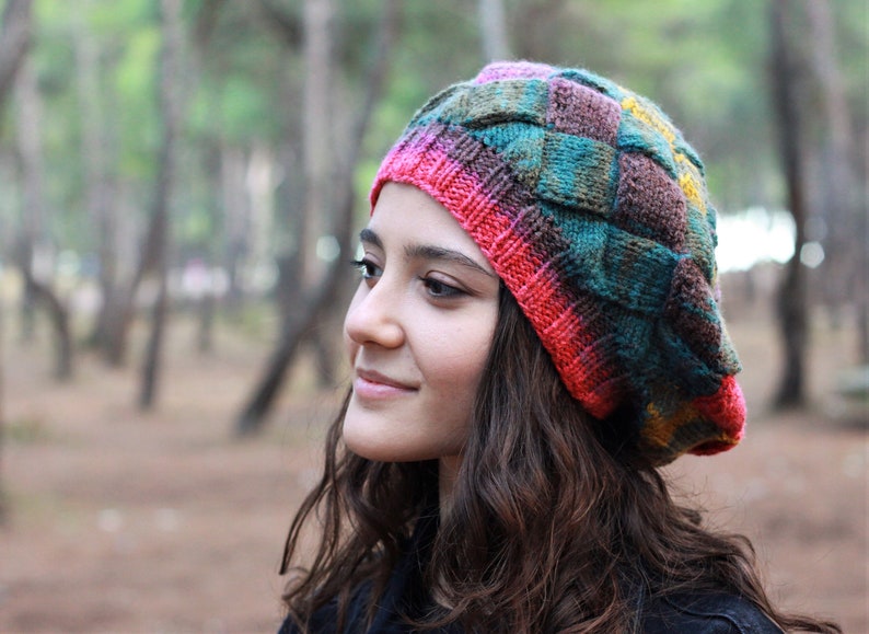 May include: A woman wearing a colourful knitted beanie with a multi-coloured pattern. The beanie is made of yarn in shades of pink, green, brown, and yellow. The woman has long brown hair and is looking off to the side.