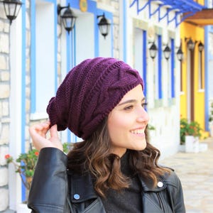 Pode incluir: Um gorro de malha roxo escuro com um design de malha entran&ccedil;ada. O chap&eacute;u &eacute; usado por uma pessoa que usa uma jaqueta de couro preta. O fundo apresenta edif&iacute;cios coloridos com detalhes em azul e amarelo.