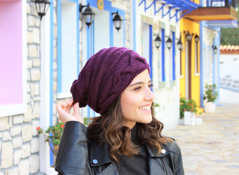 Pode incluir: Um gorro de malha roxo escuro com um design de malha entran&ccedil;ada. O chap&eacute;u &eacute; usado por uma pessoa que usa uma jaqueta de couro preta. O fundo apresenta edif&iacute;cios coloridos com detalhes em azul e amarelo.