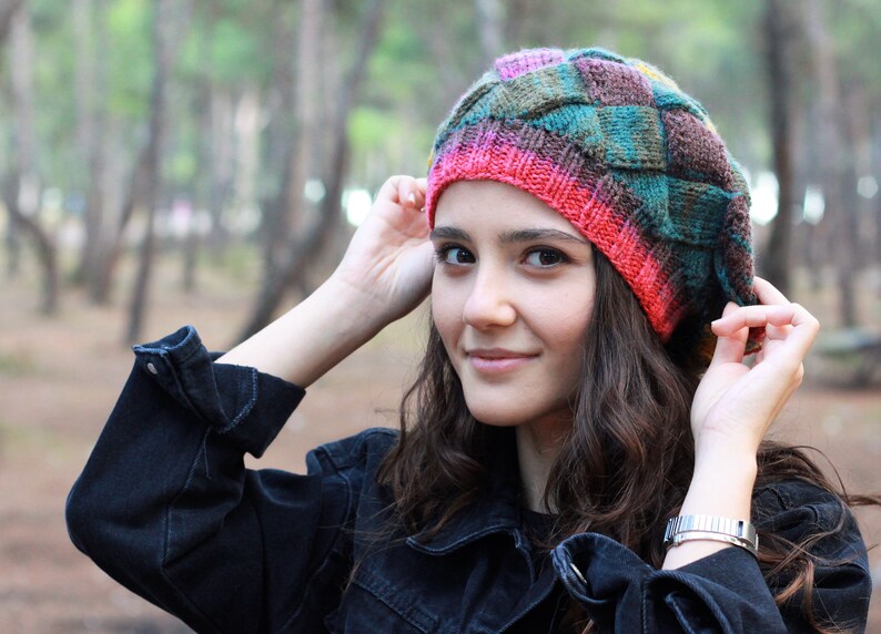May include: A woman wearing a colourful knitted beanie with a plaid pattern. The beanie is made with a mix of pink, green, brown, and blue yarn. She is wearing a dark denim jacket and has long brown hair.