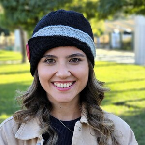 May include: A woman wearing a black and gray knitted hat with a red button. The hat has a wide brim and is angled to the side. The woman is smiling and has long brown hair.