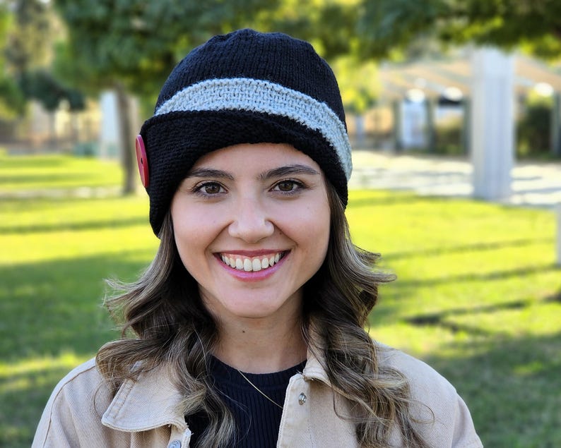 May include: A woman wearing a black and gray knitted hat with a red button. The hat has a wide brim and is angled to the side. The woman is smiling and has long brown hair.