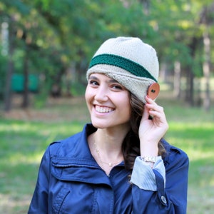 May include: A woman wearing a beige knitted hat with a green stripe and a brown button. The hat has a slouchy style and is worn tilted to the side. She is smiling and has long brown hair.