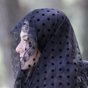 May include: A black veil with a polka dot pattern and a decorative lace trim. The veil is draped over the head and shoulders, creating a soft, elegant look. The background is blurred, focusing attention on the veil.