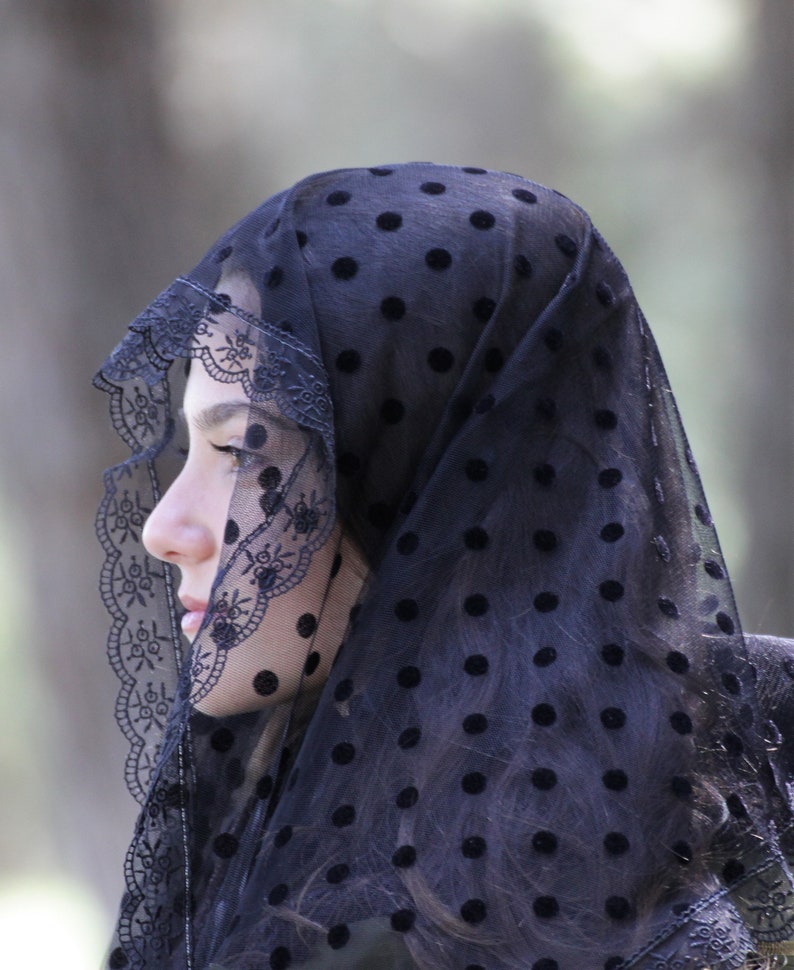 May include: A black veil with a polka dot pattern and a decorative lace trim. The veil is draped over the head and shoulders, creating a soft, elegant look. The background is blurred, focusing attention on the veil.