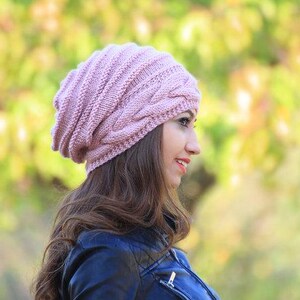 Pode incluir: Um gorro de malha rosa claro com um design de malha de cabo. O chap&eacute;u &eacute; feito de um fio macio e texturizado e tem um ajuste folgado. A pessoa est&aacute; usando uma jaqueta azul escura.