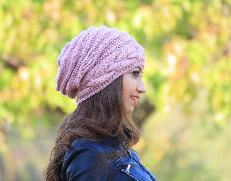 Pode incluir: Um gorro de malha rosa claro com um design de malha de cabo. O chap&eacute;u &eacute; feito de um fio macio e texturizado e tem um ajuste folgado. A pessoa est&aacute; usando uma jaqueta azul escura.
