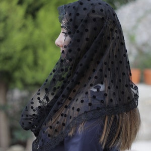 May include: Black veil with a polka dot pattern and lace trim. The veil is draped over the head and shoulders, with the wearer's profile visible. The background is blurred, with green foliage and a blue garment.
