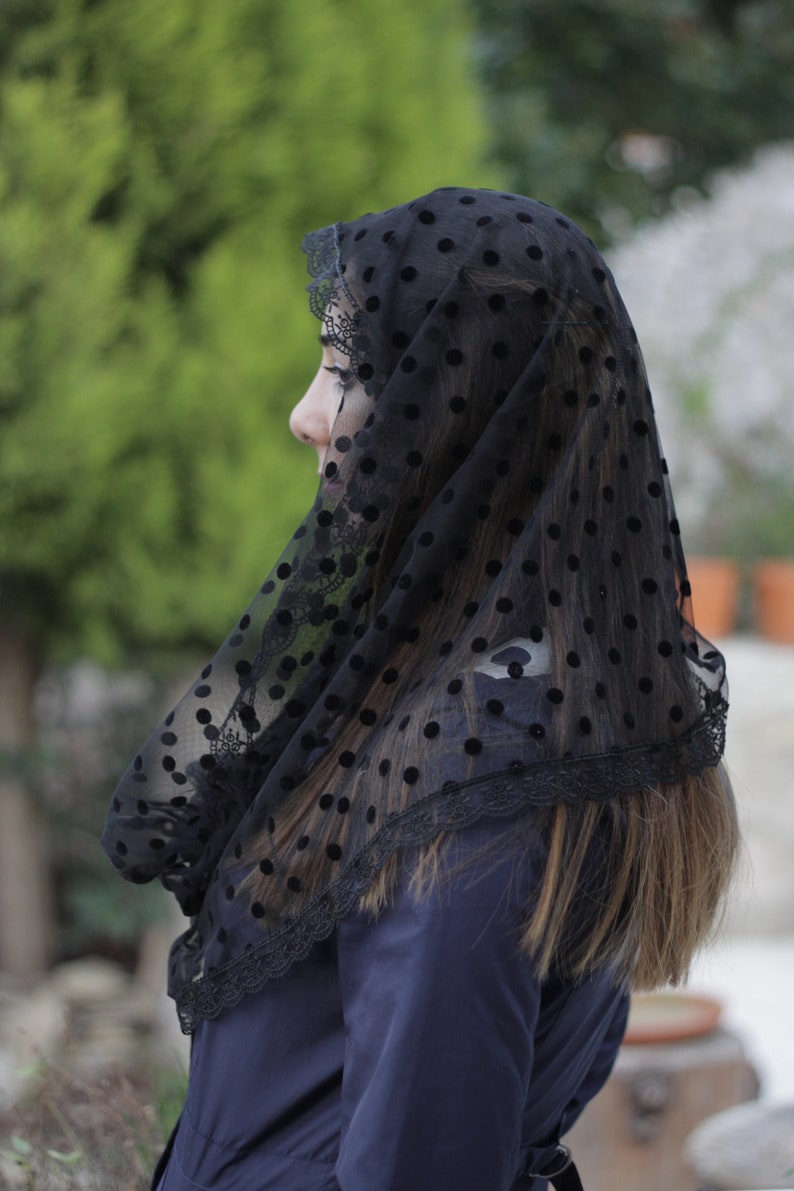 May include: Black veil with a polka dot pattern and lace trim. The veil is draped over the head and shoulders, with the wearer's profile visible. The background is blurred, with green foliage and a blue garment.