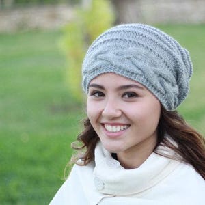 Pode incluir: Um gorro cinza claro de malha de cabo. O chap&eacute;u &eacute; feito de um fio macio e texturizado e apresenta um design cl&aacute;ssico de malha de cabo. O chap&eacute;u &eacute; usado por uma pessoa com cabelo castanho comprido e um casaco branco.