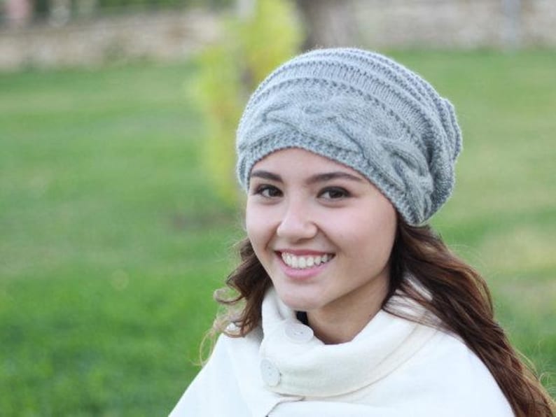 Pode incluir: Um gorro cinza claro de malha de cabo. O chap&eacute;u &eacute; feito de um fio macio e texturizado e apresenta um design cl&aacute;ssico de malha de cabo. O chap&eacute;u &eacute; usado por uma pessoa com cabelo castanho comprido e um casaco branco.