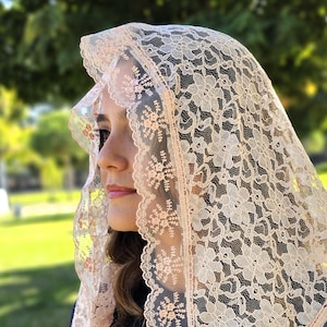 May include: A beige lace veil featuring a floral design. The veil is worn over the head and shoulders, with the face partially visible. The lace has an intricate pattern, and the colour is a soft, neutral tone.