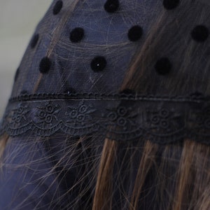 May include: Black polka dot veil with a delicate lace trim. The veil is made of sheer black fabric with evenly spaced, large, black polka dots. The lace trim features a floral design.