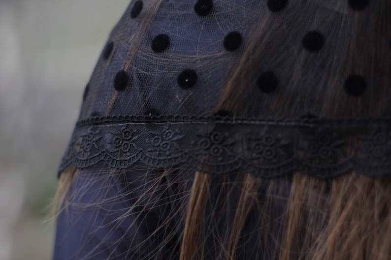 May include: Black polka dot veil with a delicate lace trim. The veil is made of sheer black fabric with evenly spaced, large, black polka dots. The lace trim features a floral design.