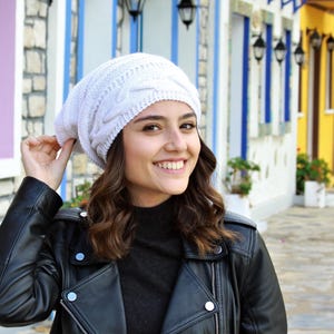 Pode incluir: Um gorro branco de malha canelada. O chap&eacute;u &eacute; feito de um material macio e texturizado. A pessoa est&aacute; usando uma jaqueta de couro preta e uma gola alta preta. O fundo apresenta edif&iacute;cios coloridos.