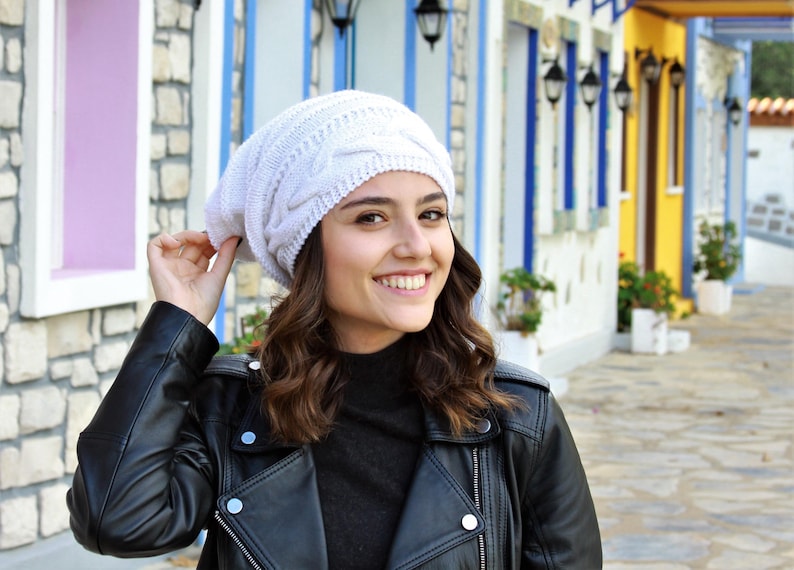 Pode incluir: Um gorro branco de malha canelada. O chap&eacute;u &eacute; feito de um material macio e texturizado. A pessoa est&aacute; usando uma jaqueta de couro preta e uma gola alta preta. O fundo apresenta edif&iacute;cios coloridos.