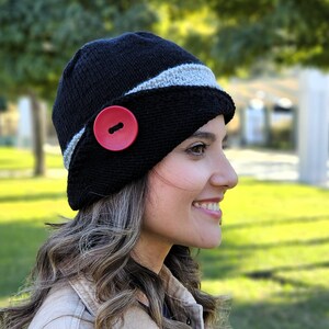 May include: A black knit hat with a gray stripe and a large red button. The hat is worn by a person with long brown hair.