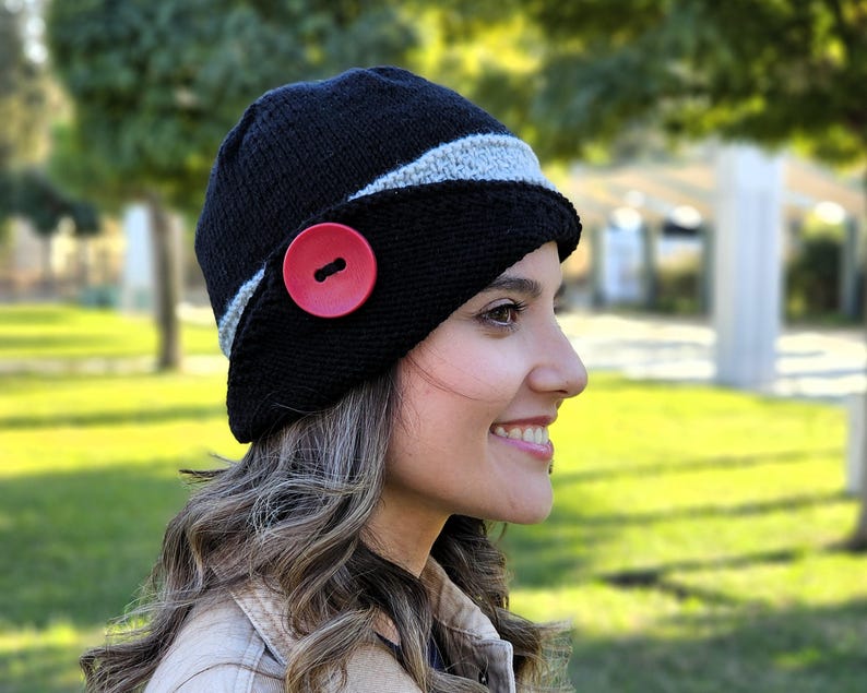 May include: A black knit hat with a gray stripe and a large red button. The hat is worn by a person with long brown hair.