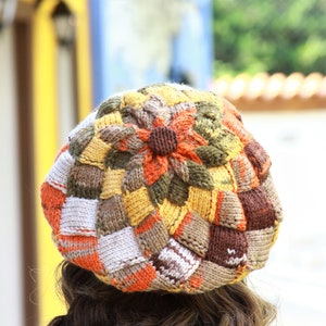 May include: A colorful knitted beret with a flower design in shades of brown, orange, white, and yellow.