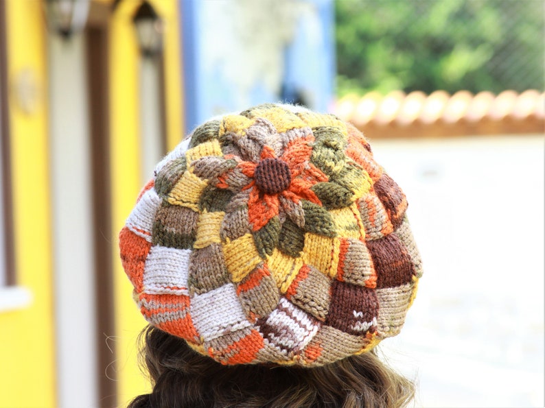 May include: A colorful knitted beret with a flower design in shades of brown, orange, white, and yellow.