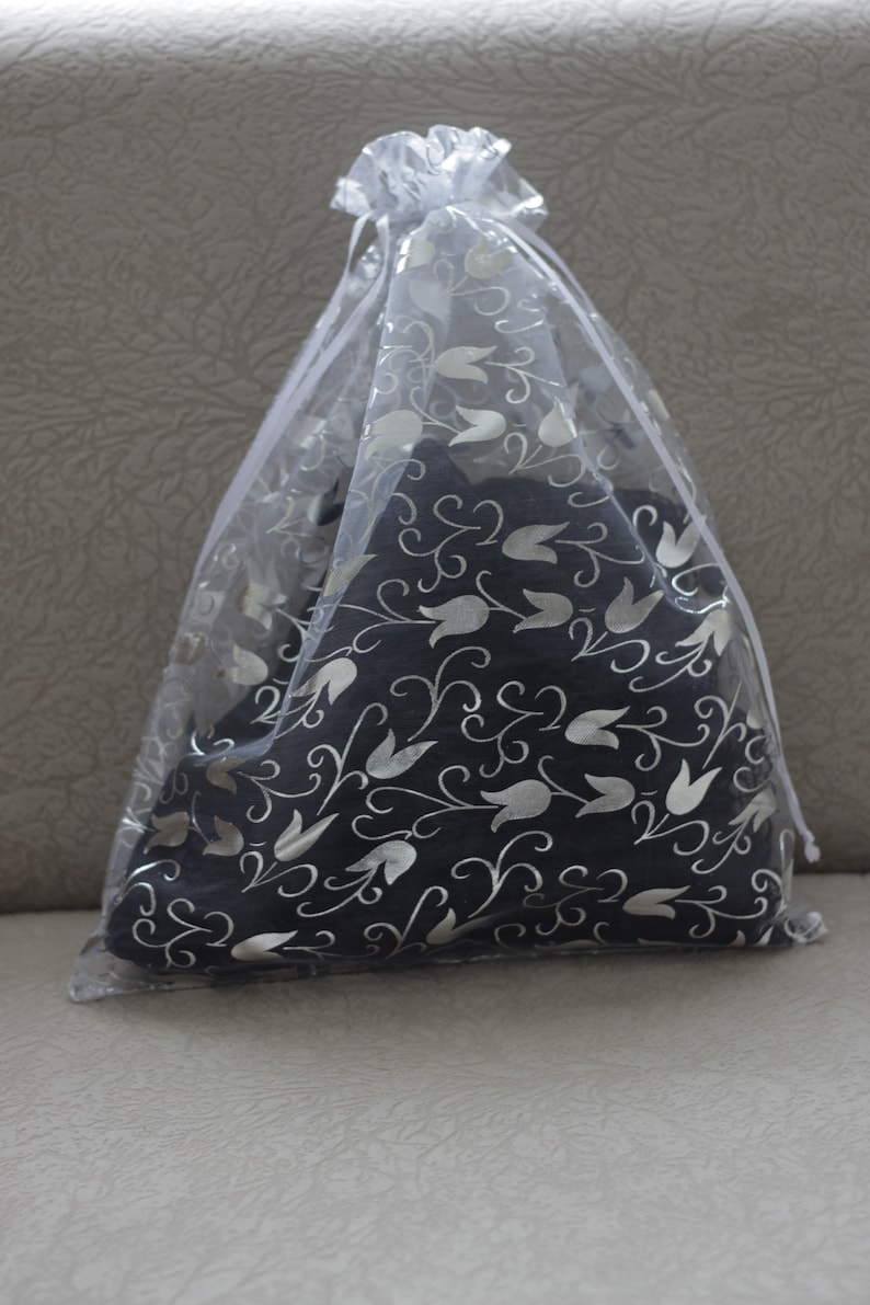 May include: A dark gray decorative pillow with a silver floral pattern is enclosed in a sheer white gift bag. The bag is gathered at the top with a white ribbon, creating a soft, elegant presentation.