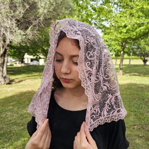 Könnte beinhalten: Ein hellrosa Spitzenschleier mit einem komplizierten Blumenmuster. Der Schleier ist über Kopf und Schultern drapiert. Die Spitze hat einen gewellten Rand. Der Hintergrund ist ein Park mit grünen Bäumen.