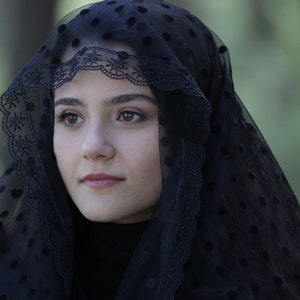 May include: A black veil with a dotted pattern and a decorative lace trim. The veil is draped over the head and shoulders. The person is wearing a black turtleneck.
